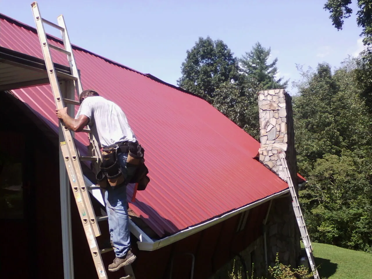 Roofer on a ladder beside a red metal roof and stone chimney — Soffit & Fascia Repair by Roofing Brothers in North Collins